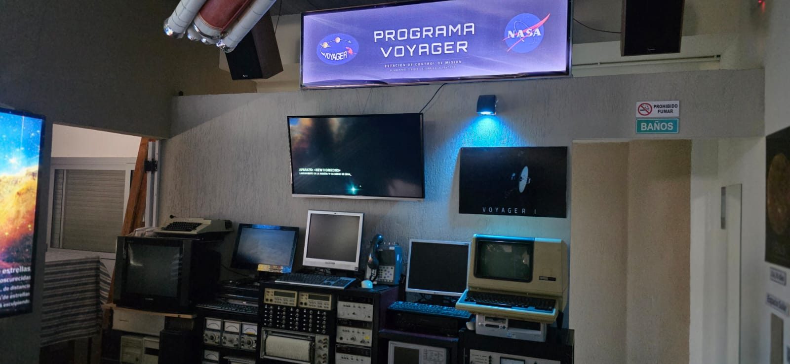 Vista de la Estación de Control de Misión del Programa Voyager en el planetario, con monitores, consolas y un cartel de la NASA